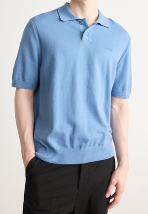 Lyseblå kortærmet polo-shirt med ribkant og diskret "BOSS"-logo, båret med sorte bukser, hænderne i lommerne.