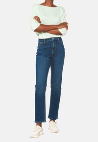 Ljusgrön, texturerad topp med vida ärmar; åtsittande mörkblå jeans; beige sneakers. Avslappnad outfit med modern, minimal design.