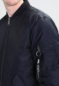 Giubbotto bomber navy con zip frontale, collo a coste e tasca con patta sulla manica. Presenta un portachiavi e un'etichetta in tessuto.