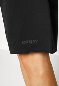 Fekete sportnadrág sima textúrával, oldalsó varrásokkal és a hemnél elhelyezett domború "Oakley" logóval.