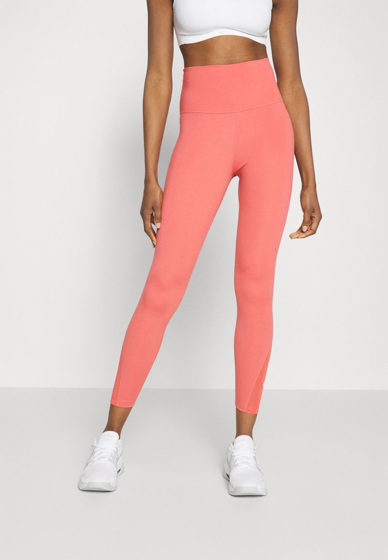 Legginsy w kolorze koralowym, z wysokim stanem, wykonane z gładkiego, elastycznego materiału. Charakteryzują się dopasowanym krojem i długością do kostek, w połączeniu z białymi butami sportowymi.