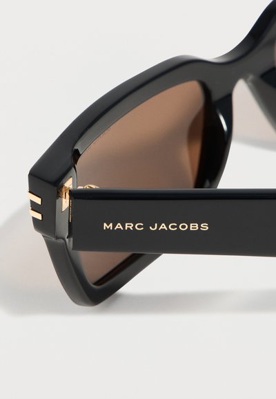Μαύρα γυαλιά ηλίου Marc Jacobs με χοντρά πλαίσια, καφέ φακούς και χρυσό λογότυπο στο μπράτσο πάνω σε ανοιχτό φόντο.