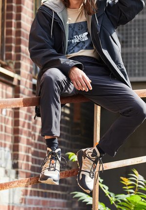 Personne portant une veste noire, un pantalon noir et des baskets beige et noir assise sur une rambarde métallique rouillée à l'extérieur près d'un mur en briques.