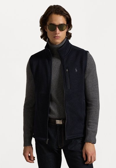 Marine blauwe gebreide vest met hoge kraag, grijze turtleneck eronder en donkere spijkerbroek. Bevat een ritsschke pocket en logo accent.