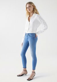 Camisa blanca de botones con detalles bordados, combinada con jeans ajustados de color azul claro y tacones negros con tiras. Diseño simple y limpio.