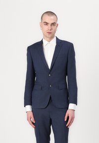 Navy blue suit with a textured fabric, featuring a two-button closure, notch lapels, and a white dress shirt underneath. Earring detail visible.