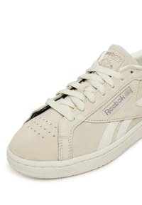 Zapatillas de ante beige con acentos en cuero blanco, punta redonda, detalles perforados en la caja del dedo y cordones planos. Presenta el logo de Reebok.