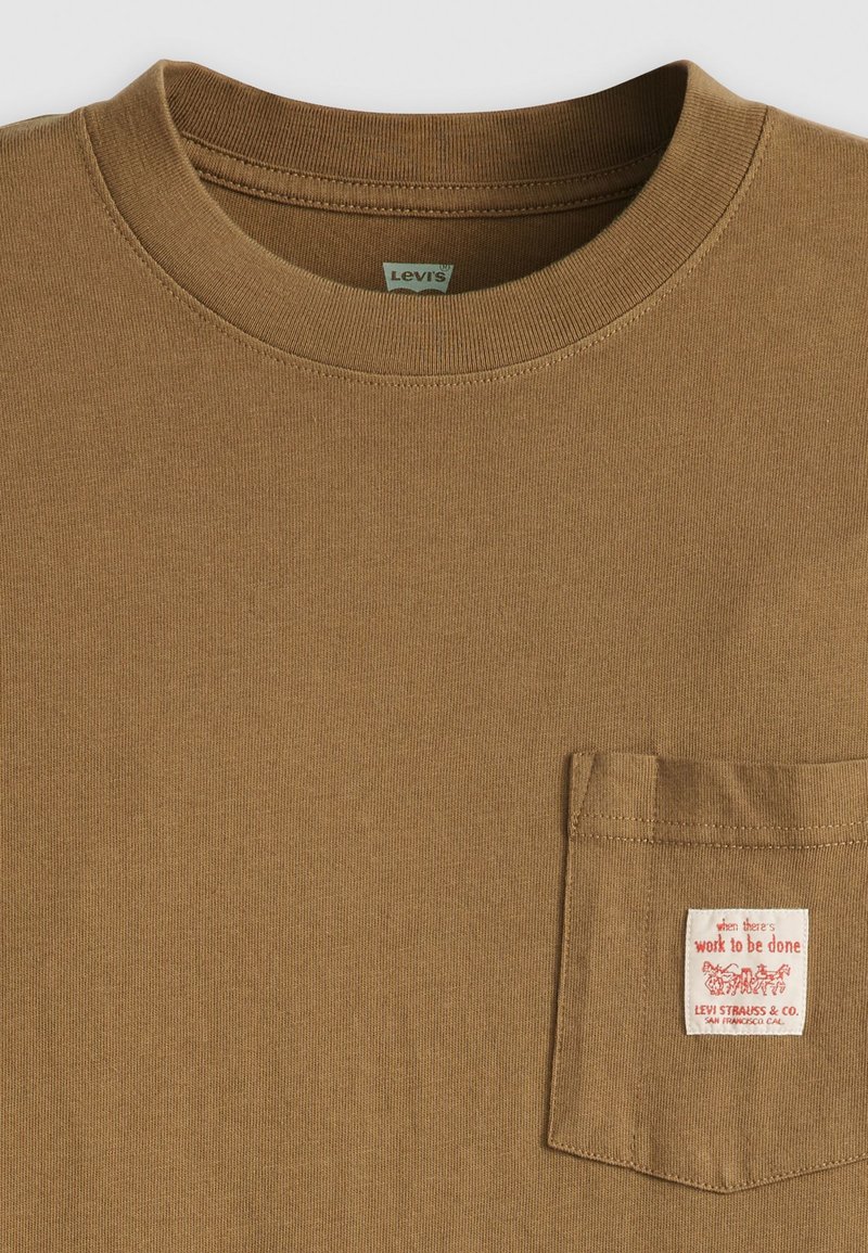 T-shirt in cotone marrone con colletto rotondo. Presenta una tasca sul petto sinistro con un'etichetta rossa e bianca. Texture liscia, design casual.