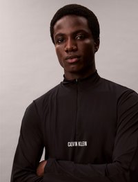 Črn pulover s pol zadrgo in dolgimi rokavi, narejen iz gladkega, lahkega materiala; ima bel logo "Calvin Klein" na prsih.