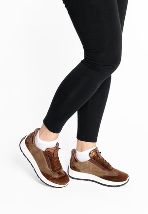 Person wearing brown suede sneakers with metallic accents, white ankle socks, and black leggings standing on a white background.