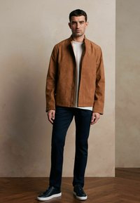 Brązowa zamszowa kurtka z kołnierzem typu stand-up, zamek błyskawiczny, i kieszeniami z przodu, zestawiona z białym T-shirtem i granatowymi spodniami oraz ciemnymi sneakersami.