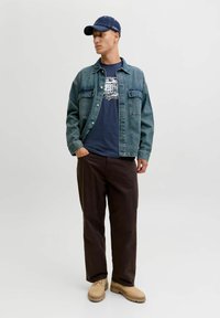 Joven vestido con una chaqueta de mezclilla, camiseta gráfica azul marino, pantalones marrón oscuro, botas beige y una gorra de béisbol azul, de pie contra un fondo liso.