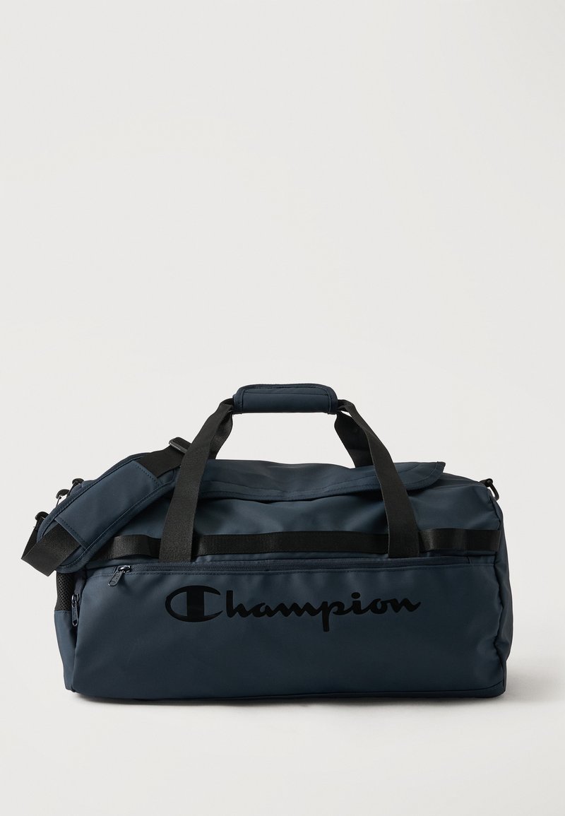 Μεγάλη ναυτικό μπλε τσάντα Champion τύπου duffel με μαύρες λαβές, ιμάντα ώμου και μπροστινή τσέπη με φερμουάρ σε απλό ανοιχτόχρωμο φόντο.