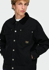 Črna denim jakna z ovratnikom iz kordurovega blaga, zapenjanjem na pritisne gumbe, dvema žepoma na prsih in majhno logotipno značko na levem žepu.