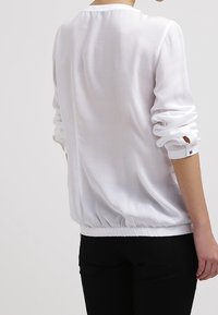 Vit, lätt blus med långa ärmar, rynkade manschetter och rundad nederkant. Slät textur och enkel design, lämplig för lager-på-lager.