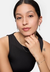 Bijoux en argent avec chaînes, incluant des boucles d'oreilles pendantes et des bracelets. Le modèle porte un top sans manches noir, mettant en valeur une peau lisse et des tatouages.