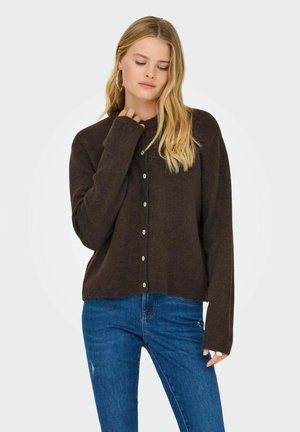Jeune femme aux cheveux blonds portant un cardigan marron boutonné et un jean bleu, posant avec une main près du cou et les yeux fermés.