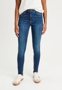 Jean skinny en denim bleu foncé, avec un design ajusté, taille mi-haute et couture discrète. Associé à des baskets blanches et beiges.