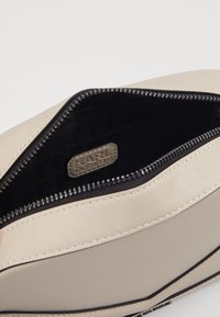 Beżowa kieszonka na zamek z teksturowanym płóciennym zewnętrzem i gładkim wnętrzem, wyposażona w metkę z logo oraz czarną podszewkę.