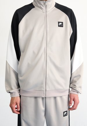 Person iført en beige, sort og hvid Nike Air lynlåstrøje og matchende beige bukser med Nike Air logo patches.