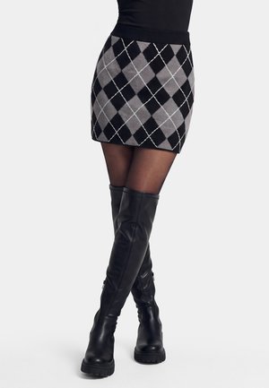 Kobieta w mini spódniczce z czarno-szarym wzorem argyle, przezroczystych czarnych rajstopach i czarnych skórzanych botkach za kolano z grubą podeszwą.