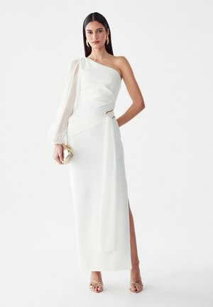 Femme portant une robe blanche asymétrique avec une manche longue transparente, tenant une pochette dorée et portant des sandales et des boucles d'oreilles dorées.