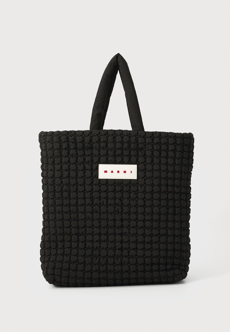 Fekete steppelt táska párnázott fülekkel, amelyen egy téglalap alakú fehér címke található piros felirattal. Puha, texturált felület.