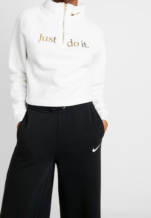 Persoon draagt een witte sweat met rits en gouden "Just do it." tekst en zwarte broek met een wit Nike Swoosh-logo, handen in de zakken.