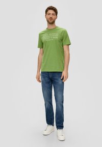 Zeleni pamučni T-shirt s natpisom "OLIVER" ispisanim bijelom bojom. Okrugli izrez, kratki rukavi, uparen s plavim trapericama i bijelim tenisicama.