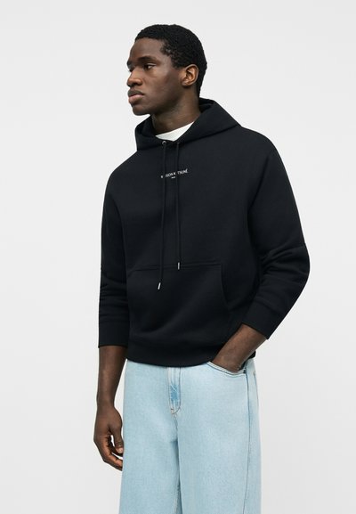 Νεαρός άνδρας που φοράει μαύρο φούτερ με κουκούλα και τσέπη μπροστά και ανοιχτόχρωμο τζιν, στέκεται με το ένα χέρι στην τσέπη, κοιτάζοντας αριστερά.