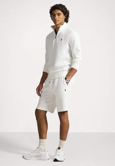 Giovane uomo che indossa una felpa beige a metà zip, pantaloni corti coordinati, calze beige e scarpe da ginnastica bianche, in piedi con una mano in tasca.