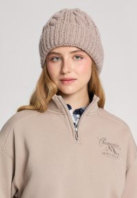 Beżowa czapka wykonana na drutach z prążkowanym wzorem i splotem na górze, zestawiona z beżowym polarowym zamek, na którym znajduje się haftowane logo.