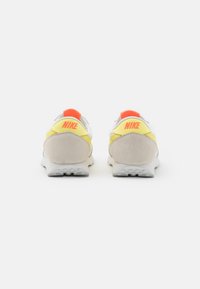 Tenisky Nike s bílou síťovinou a koženým svrškem, žlutými a oranžovými akcenty a texturovanou šedou semišovou patou. Mezi detaily se nachází viditelné logo.