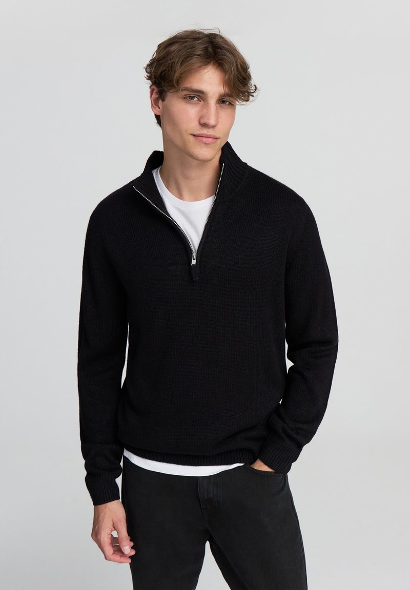 Maglione nero a maglia con colletto alto e chiusura a zip, tessuto testurizzato, vestibilità rilassata, indossato sopra una maglietta bianca, abbinato a pantaloni neri.