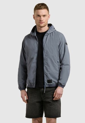 Muž v svetlomodrej bunde s kapucňou cez čierne tričko a tmavosivé šortky stojí pred obyčajným svetlým pozadím.