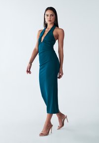 Teal halterneck-mekko, jossa on tiukka siluetti, rypytys vyötäröllä ja etuosan avausyksityiskohta, yhdistettynä beigeihin remmikenkiin.