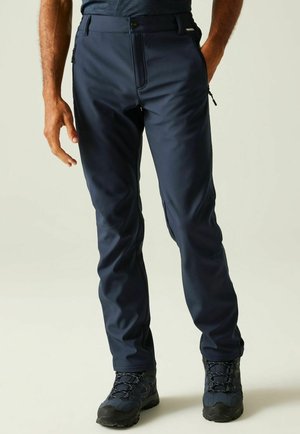 Man draagt marineblauwe outdoorbroek met ritssluitingen aan de zijzakken en zwarte wandelschoenen, staand tegen een effen achtergrond.