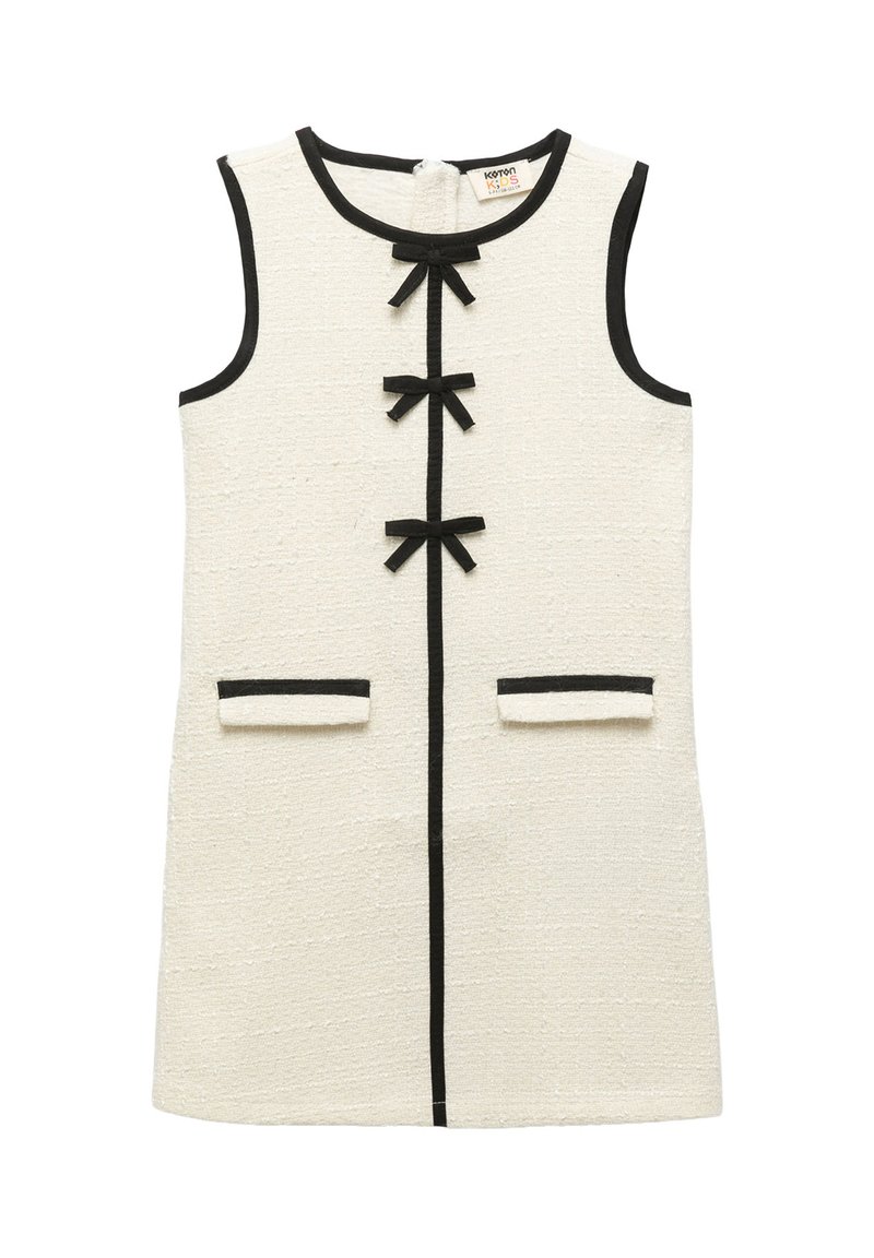 Bezramionowa sukienka w kolorze kremowym o fakturowanej powierzchni z czarnymi wykończeniami, trzy czarne kokardy na przodzie i dwie przednie kieszenie z czarnymi brzegami.