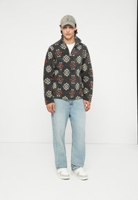 Fleece-pullover med geometrisk mönster i mörkgrått, rött och krämfärgat; har en fjärdedels dragkedja, mjuk textur och matchas med ljusblå jeans.