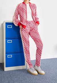 Röd och vit mönstrad träningsdräkt med bomberjacka och åtsittande byxor, ihopparad med grå sneakers, framför ett blått arkivskåp.