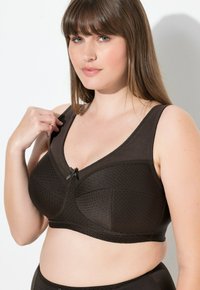 Kobieta o jasnej karnacji i brązowych włosach, ubrana w ciemnobrązowy teksturowany biustonosz z małą kokardką, poprawiająca ramiączko na tle białej ściany.