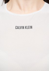 Κοντινό πλάνο ενός ατόμου που φοράει λευκό πουκάμισο από δίχτυ με την ένδειξη "CALVIN KLEIN" σε μαύρα γράμματα στο στήθος.