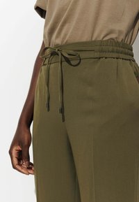 Pantalon vert olive avec une taille élastique et un cordon de serrage noué, fait d'un tissu lisse, doté de poches latérales et d'une coupe décontractée.