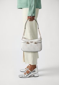 Bolso blanco con estampado, con herrajes dorados y ribete negro, sostenido con una mano. También son visibles zapatos de tacón bloque plateados y una blusa verde clara.