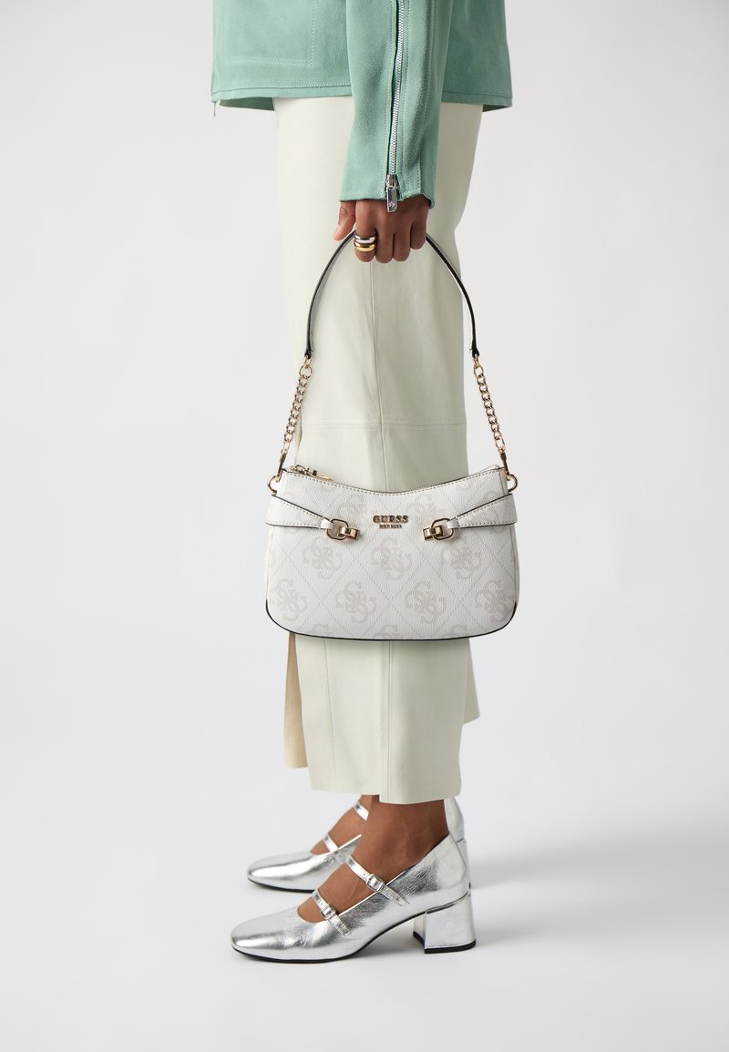 Bolso blanco con estampado, con herrajes dorados y ribete negro, sostenido con una mano. También son visibles zapatos de tacón bloque plateados y una blusa verde clara.