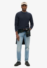 Marinefarbener Strickpullover, hellblaue Jeans, grüne Flanellhemd um die Taille gebunden, beiger Hut, schwarze Schuhe mit strukturierter Oberfläche, kurze Ärmel.