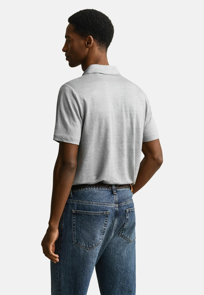 Mann von hinten gesehen, trägt ein hellgraues kurzärmeliges Polohemd und blaue Jeans vor einem weißen, einfarbigen Hintergrund.