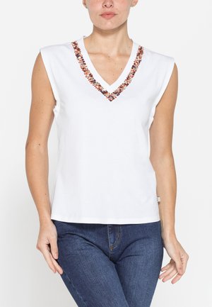 Donna che indossa un top bianco senza maniche con scollo a V decorato con perline marroni e arancioni, abbinato a jeans blu.