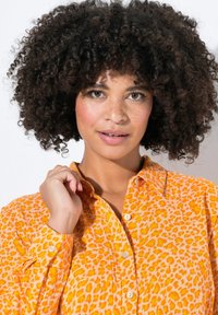 Oranžová košile s leopardím potiskem, s knoflíkovým zapínáním a límcem, vyrobená z texturované látky. Model se dotýká límce.