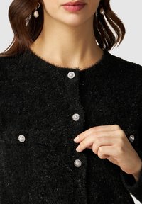 Svart, fluffig cardigan med runda pärlknappar. Har en rund halsringning och mjuk textur. Hand som vilar på den främre knappen.